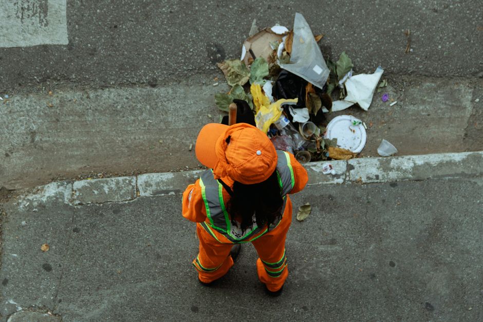 A waste collection worker dressed in green high-visibility work uniform with reflective orange stripes is standing on the pavement beside an open refuse collection vehicle, which is parked on a reside