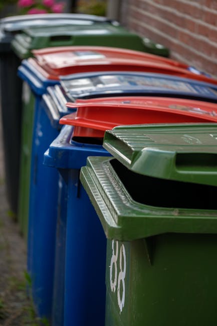 A row of multiple large wheeled rubbish bins located along a pavement adjacent to a brick wall. The bins are predominantly blue with green, red, and dark green lids, each having a raised, textured sur
