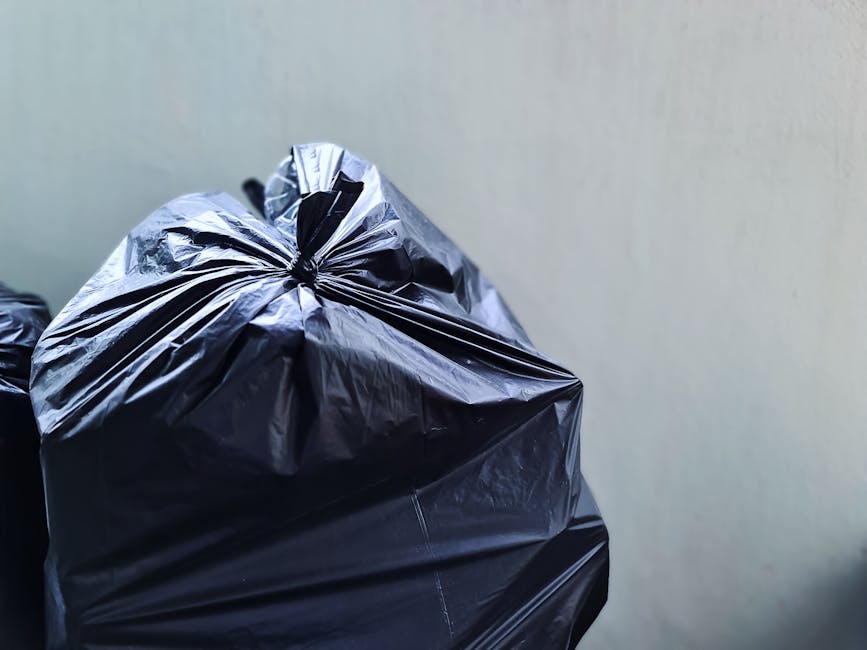 Close-up image of two large black plastic rubbish bags filled with waste, tightly knotted at the top, with a slightly glossy surface reflecting light. The bags are stacked against a plain, light-color